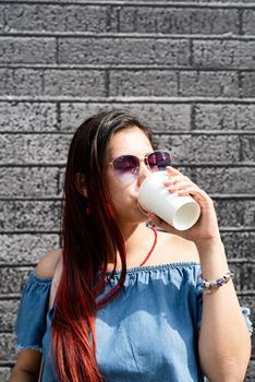 coffee to go. stylish young woman wearing jeans shirt, sunglasses and bag, drinking coffee to go at street, black brick wall background