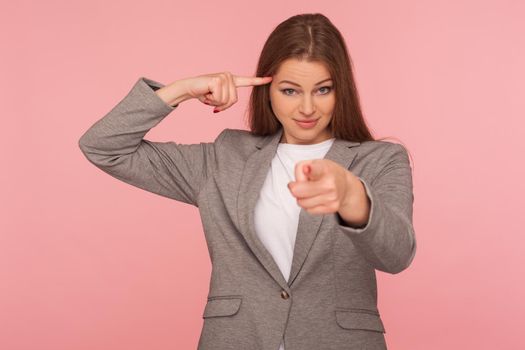 You are insane, out of mind. Portrait of young woman in business suit showing stupid gesture and pointing to camera, displeased with your reckless silly joke, dumb idea. indoor studio shot, isolated