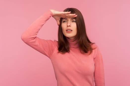 Curious brunette woman in pink sweater looking far away with hand over head, trying to see something, bad vision. Indoor studio shot isolated on pink background