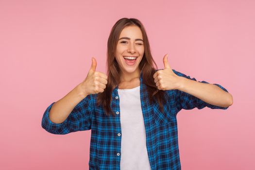Well done. Portrait of enthusiastic girl in checkered shirt standing with double thumbs up and smiling broadly at camera, pleased with result, rating excellent. studio shot isolated on pink background
