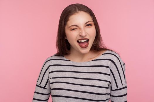 Portrait of coquettish playful beautiful woman in striped sweatshirt flirting to camera, blinking eye and looking carefree, cheering up with wink. indoor studio shot isolated on pink background