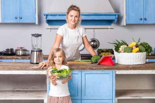 Young beautiful mother standing at table while her daughter holding bowl of salad with fresh vegetables, girls smiling posing on camera in modern kitchen, showing cooked vegetarian food, healthy diet