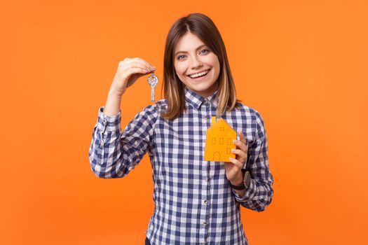 Portrait of home owner, brunette woman with charming smile wearing checkered shirt standing holding paper house and keys, estate purchase or mortgage. indoor studio shot isolated on orange background