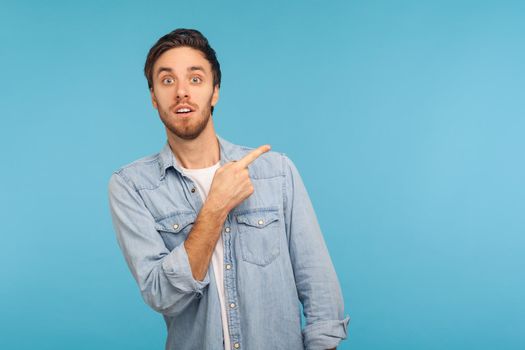 Wow, look at ads here. Portrait of surprised shocked man in denim shirt pointing aside, showing blank copy space for idea presentation, commercial text. indoor studio shot isolated on blue background