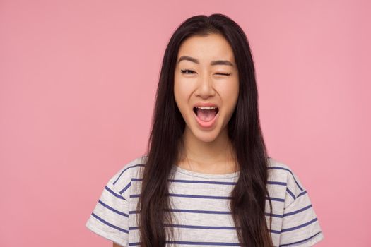 Portrait of playful girl with long hair winking at camera and smiling broadly, excited about acquaintance, having some cunning idea, blinking eye. indoor studio shot isolated on pink background