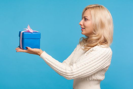 Side view beautiful woman with curly blond hair holding blue gift box in hands, making present, donation, charity concept. Indoor studio shot isolated on blue background