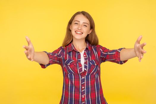 Come into my arms. Portrait of sincere happy ginger girl in shirt holding hands wide open to embrace, giving free hugs, welcoming with toothy smile. indoor studio shot isolated on yellow background