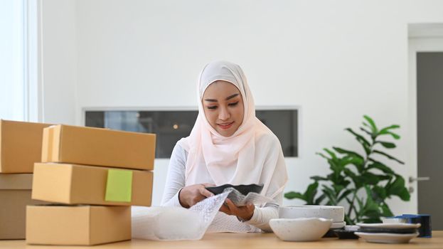 Muslim woman business owner packing product, preparing parcel for shipping to costumer. Online selling, e-commerce concept.