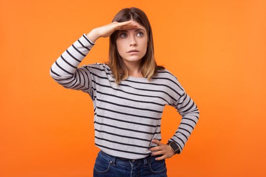 Portrait of concentrated curious young woman with brown hair in long sleeve striped shirt standing looking far away with hand above eyes, amazed stare. indoor studio shot isolated on orange background