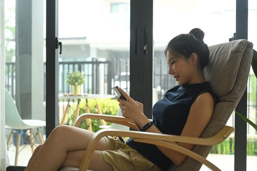 Young asian woman sitting in comfortable armchair at home and using mobile phone.