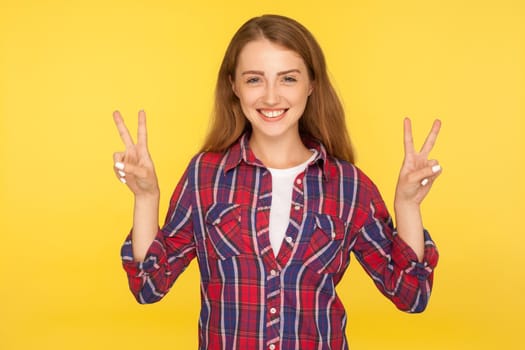 Portrait of happy successful ginger girl in checkered shirt doing victory gesture and smiling, confident in winning, showing peace sign with fingers. indoor studio shot isolated on yellow background