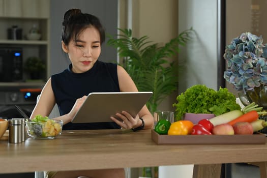 Young woman sitting in home kitchen and reading online recipe on digital tablet.