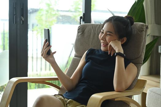 Overjoyed woman resting in comfortable armchair and making video call on smart phone.