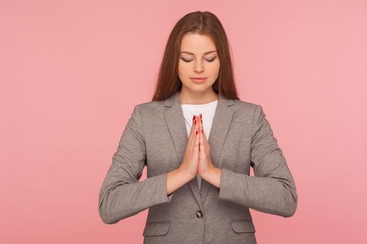Yoga and mind control. Portrait of young woman in business suit meditating with prayer hand gesture, looking calm pacified, relaxation and harmony. indoor studio shot isolated on pink background