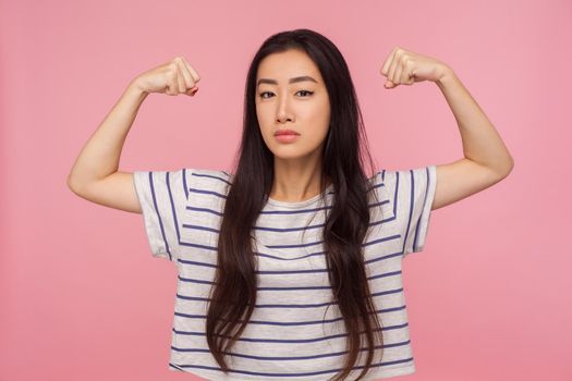 I have power, Portrait of successful bossy girl with brunette hair in striped t-shirt raising hands showing biceps, feeling strength and confidence. indoor studio shot isolated on pink background