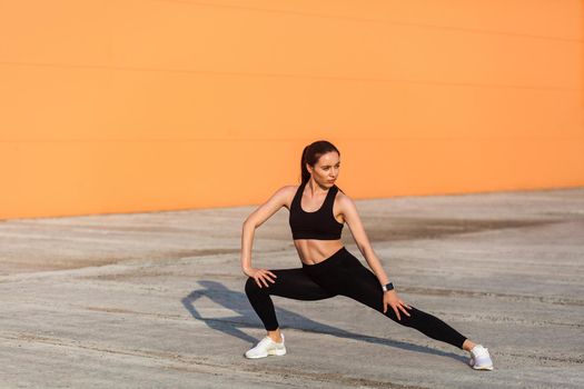 Slim young pretty woman in tight sportswear, black pants and top, practicing outdoor, doing stretching workouts for better flexibility, lower-body exercise. Health care concept, sport activity