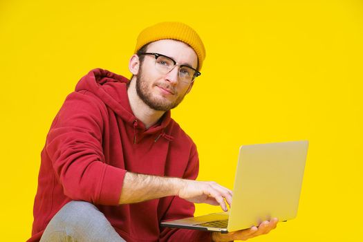 Young hipster man sitting on the floor holding laptop in red hoodie studying or working. Smart freelancer caucasian man with laptop isolated on yellow background.