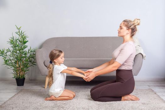 Young mother and little daughter keeping eyes closed sitting on floor, holding hands together and practicing yoga at home, doing exercise breath technique, meditating in room, mindfulness and harmony