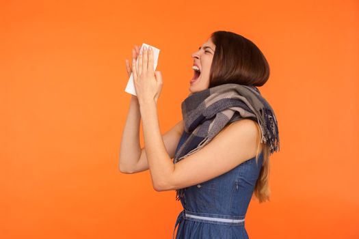 Influenza disease. Side view of flu-sick woman wrapped in scarf standing with wide open mouth and sneezing loudly in tissue, viral infection symptoms. indoor studio shot isolated on orange background