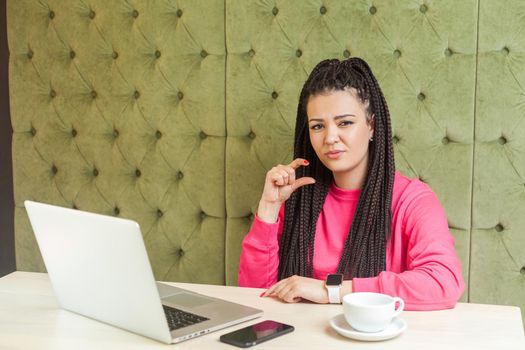 Portrait of attractive young girl with black dreadlocks hairstyle in pink blouse are sitting in cafe, working and showing little size with finger, looking at camera. Indoor, lifestyle