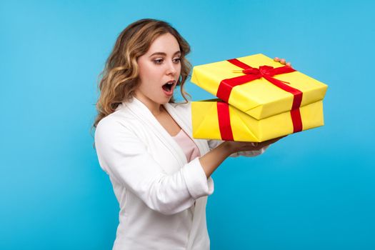 Portrait of adorable surprised woman with wavy hair in white jacket peeking with curious amazed look into gift box, unpacking interesting present. indoor studio shot isolated on blue background