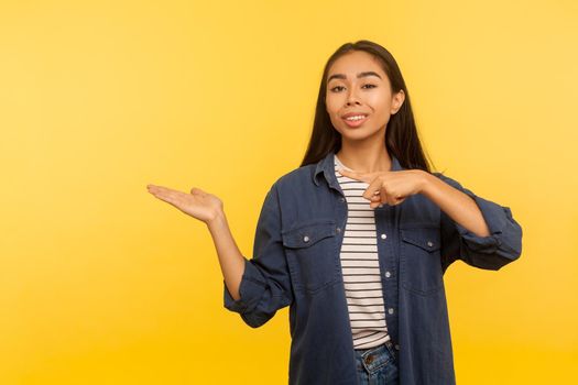 Attention, nice product advertise. Portrait of happy girl in denim shirt holding copy space with palm, pointing at empty place for commercial image. indoor studio shot isolated on yellow background
