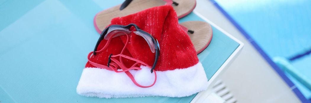 Hat of Santa Claus, swimming slippers on sunbed. Holidays near sea pool for Christmas holidays and relaxation, leisure, tourism concept.