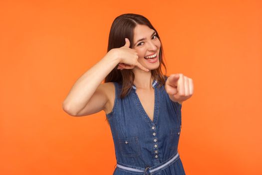 Hey you, call me. Happy playful cute brunette woman in denim dress holding arm shaped in telephone gesture and pointing to camera, looking flirtatious. indoor studio shot isolated on orange background