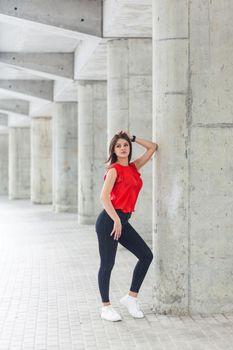 beautiful woman in red blouse and jeans posing while leaning on gray column. outdoor shot.