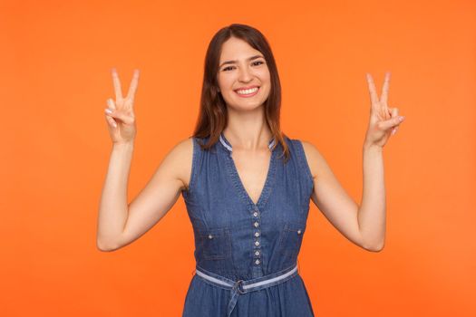 Peace and victory to you. Friendly cheerful brunette woman in denim dress making double v gesture and smiling at camera, showing sign of success, triumph. studio shot isolated on orange background