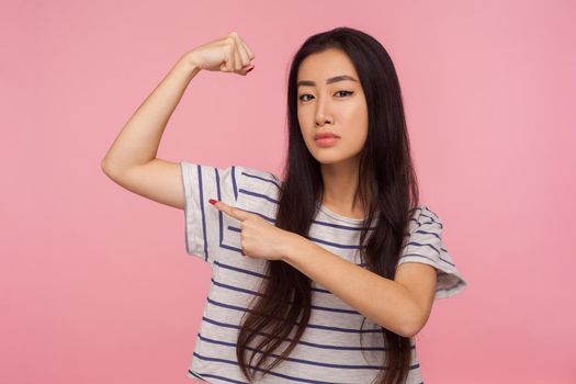 Look, I'm strong, Portrait of successful attractive girl with brunette hair in striped t-shirt showing biceps on raised hand, feeling power and confidence. studio shot isolated on pink background