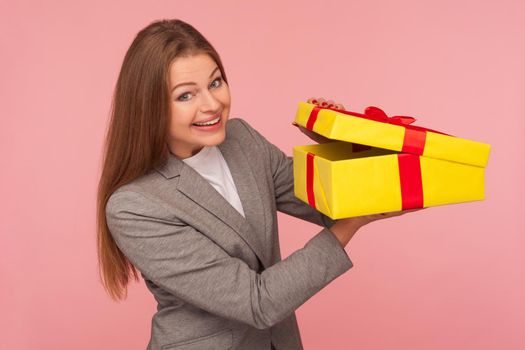 Portrait of attractive happy businesswoman in suit jacket opening gift box and looking excitedly in anticipation of surprise, unwrapping birthday present. studio shot isolated on pink background