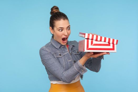 Astonished curious woman with hair bun in denim jacket unpacking opening small carton box, looking inside with surprised amazed expression, shocked by gift. studio shot isolated on blue background