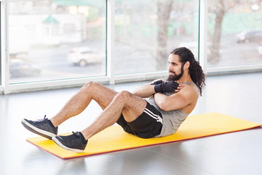 Portrait of young adult muscular built athlete with long curly hair working out in gym, lying on mat and swinging press with crossed arms on chest and bent legs. indoor, looking away,window background