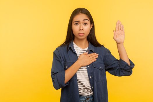 I swear. Portrait of honest girl in denim shirt taking sacred oath, making solemn vow in ceremonial tradition, affirmation of promise and devotion. indoor studio shot isolated on yellow background
