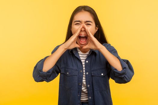 Attention, everyone listen. Portrait of angry girl in denim shirt shouting into hands shaped in megaphone, announcing negative message, loud voice. indoor studio shot isolated on yellow background