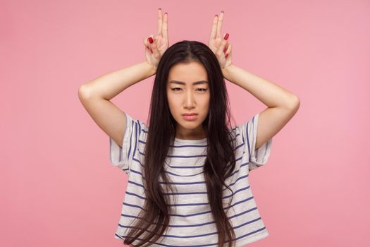Portrait of irritated girl in striped t-shirt threatening with bull horn gesture, looking at camera with hostile suspicious expression, ready to attack. indoor studio shot isolated on pink background