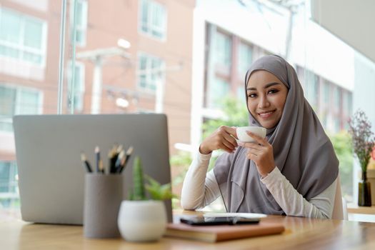 Attractive muslin woman working in office with laptop computer, hand holding cup of coffee