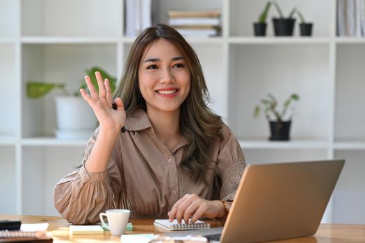 Friendly asian woman office worker greeting colleagues while working with computer laptop.
