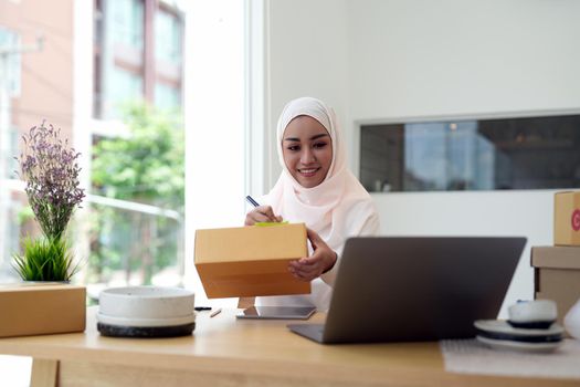 Happy young muslim woman owner writing address on cardboard box, Young Owner Woman Start up for Business Online. People with online shopping SME entrepreneur or freelance working concept