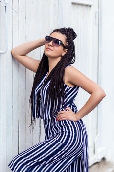 Portrait of young brunette woman with black afro american dreadlocks hairstyle in striped dress and sunglasses, standing, posing and looking away and smiling. indoor studio shot on white wall.