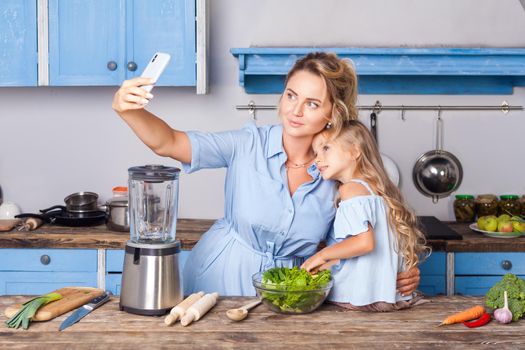 Charming woman and little girl taking selfie in modern kitchen, holding bowl of vegetable salad and posting photo in social network using cell phone, food blog about healthy nutrition, vegetarian diet