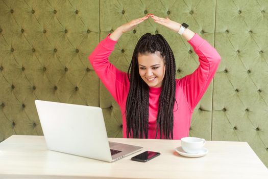 I'm in safety. Portrait of childish young girl with black dreadlocks hairstyle in pink blouse are sitting in office and holding hand upper hand, making roof with hands, looking at laptop, toothy smile