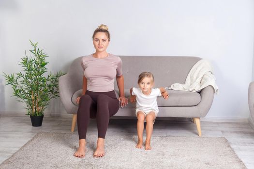 Young pretty woman and her cute daughter squatting leaning hands around on sofa, happy family doing gymnastics exercise together at home, fitness training with child. body care, healthy lifestyle