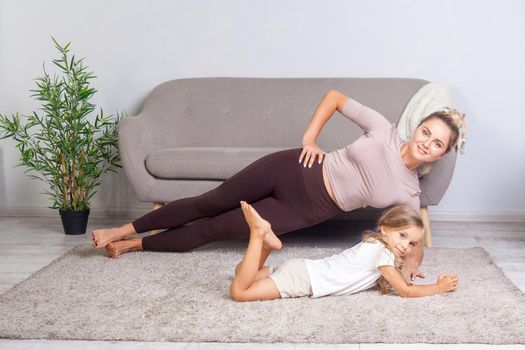 Adorable child lying near young attractive mother practicing yoga, side plank pose, happy family doing gymnastics exercise together, fitness training with child at home. body care, healthy lifestyle