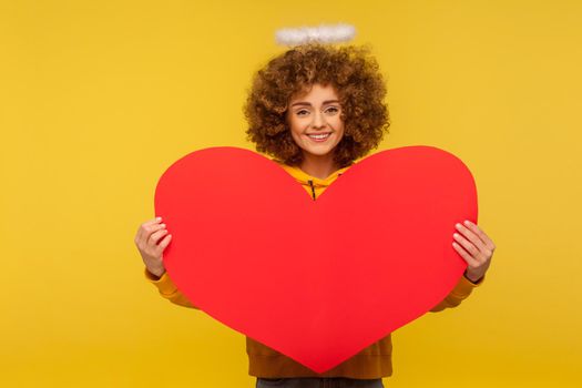 Love you, Happy Valentine's day. Portrait of attractive curly-haired woman with saint nimbus holding big red heart and smiling to camera, demonstrating romantic feelings. indoor studio shot isolated