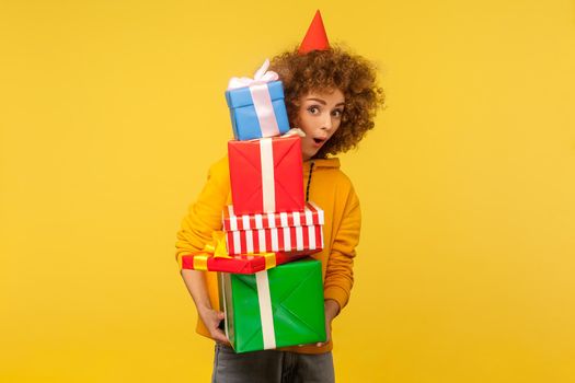Surprised emotions from big mountain of gifts. Portrait of amazed curly-haired woman with party cone holding many present boxes and looking with shocked expression. indoor studio shot, isolated