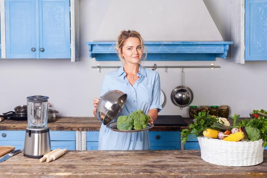 Wonderful young woman holding broccoli on tray under cloche and looking at camera, household showing cooked vegetables, healthy vegetarian food, vegan nutrition and vitamins. indoor, modern kitchen