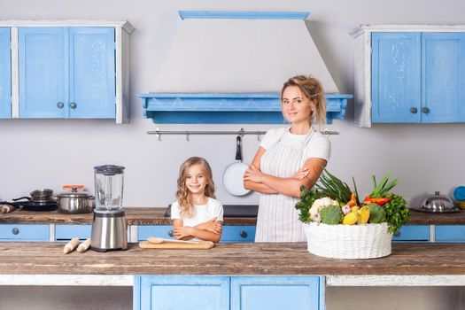 Happy beautiful mother in apron and little daughter standing with crossed hands in kitchen with modern furniture, basket of fresh green vegetables on table, eco-friendly vegetarian food, nutrition