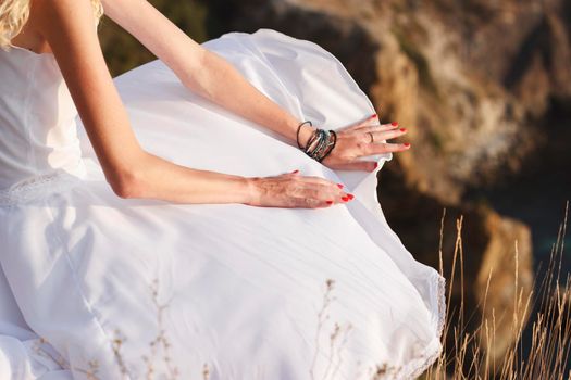 The girl holds the developing dress in the wind with her hands. Close-up of hands on a white dress. A girl is sitting on a cliff during sunset. Manicure with red varnish.